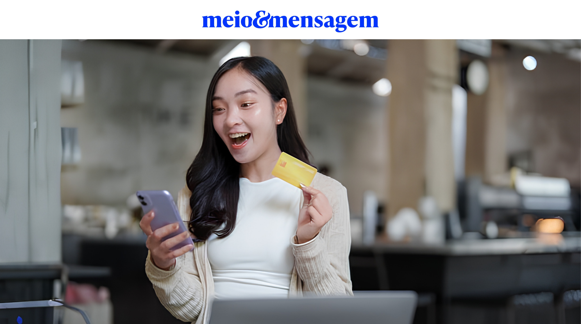 Jovem sorrindo ao olhar para o celular com um cartão de crédito na mão